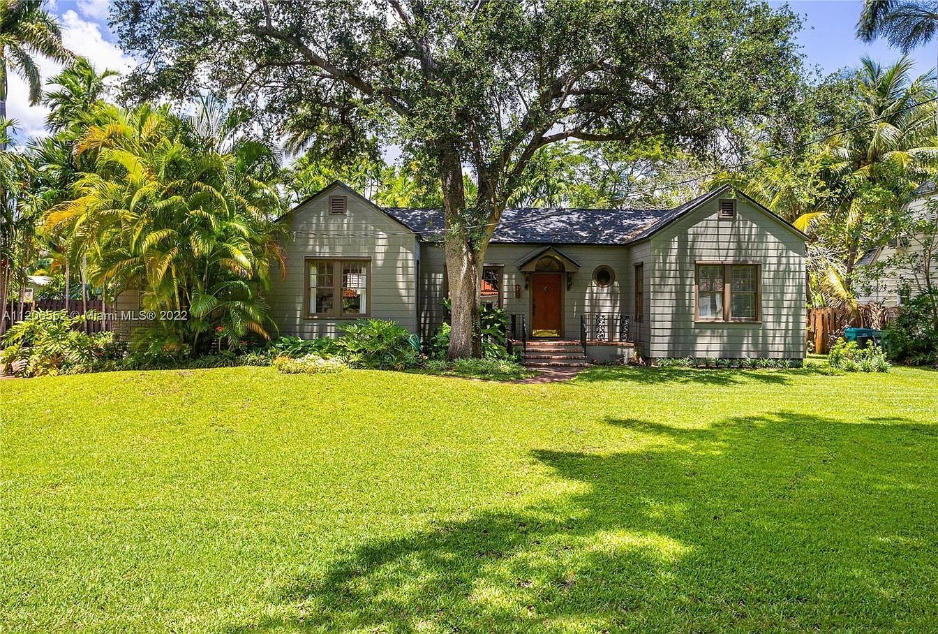 Front of 625 NE 115th St: a gray one-story cottage behind a wide lawn and a mature oak